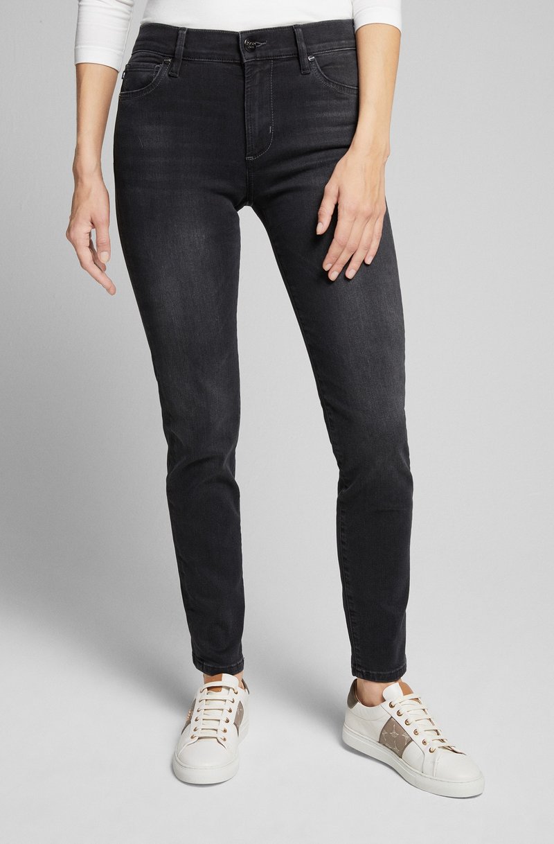 Schwarze Denim-Jeans mit Slim-Fit, ausgestattet mit vorderen Taschen, Gürtelschlaufen und einem Knopfverschluss. Kombiniert mit weißen Sneakers und einem hellen Oberteil.