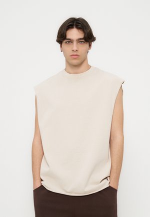 Giovane uomo con capelli castani ondulati che indossa una maglietta beige senza maniche e pantaloni scuri, in piedi davanti a uno sfondo bianco semplice.