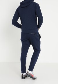 Felpa blu navy e pantaloni jogger coordinati in materiale morbido. Presenta un cappuccio con cordino e una vestibilità standard. Le calzature hanno accenti grigi e rossi.