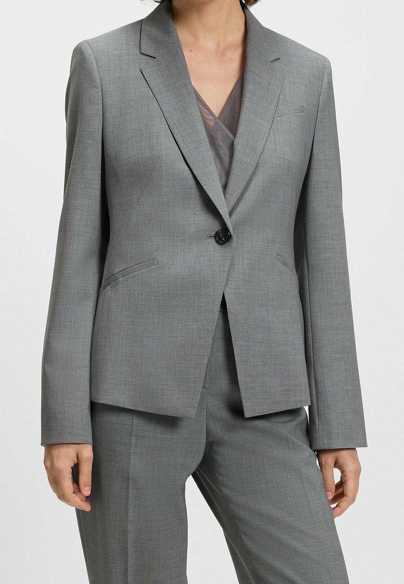 Femme portant un tailleur gris sur mesure avec un blazer à un bouton et un pantalon assorti, debout sur un fond uni.