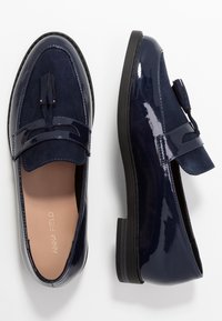 Mørkeblå loafers med blank patentering og suède-detaljer, med en dusk detalje og en lav, teksturert gummisåle.