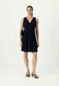  vestido sem mangas azul marinho com decote em V, cintura justinha e saia evasê. Usado com sandálias pretas de tiras e uma bolsa pequena. Tecido suave.