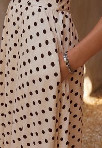 Abito beige a pois con pois marrone scuro, caratterizzato da un design plissettato e tasche. Un bracciale d'argento al polso aggiunge un accento metallico.