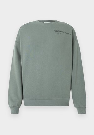 Sweat-shirt vert poussiéreux à manches longues avec un petit texte noir indiquant "Carhartt Work In Progress (Detroit) 1989 A.C 313" sur la partie supérieure gauche de la poitrine.