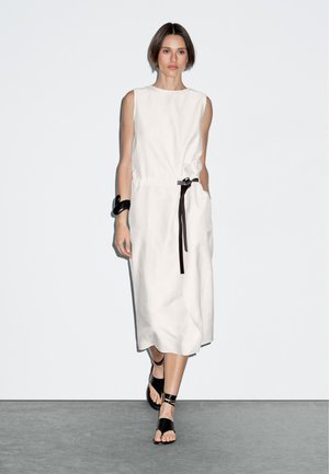 Robe blanche sans manches avec une taille cintrée et une ceinture noire, offrant une coupe ample. Accordée avec des sandales noires à brides sur un sol gris.