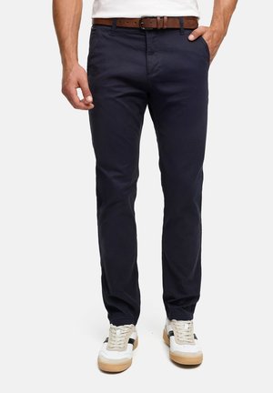 Man die marineblauwe slim-fit broek, witte sneakers met zwarte strepen, een bruine leren riem en een wit overhemd draagt, met handen bij de zakken staand.