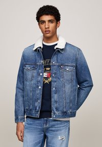 Giacca di jeans con colletto in shearling bianco, dotata di chiusure a bottoni, tasche sul petto e una lavatura azzurro chiaro; indossata sopra un maglione blu scuro.
