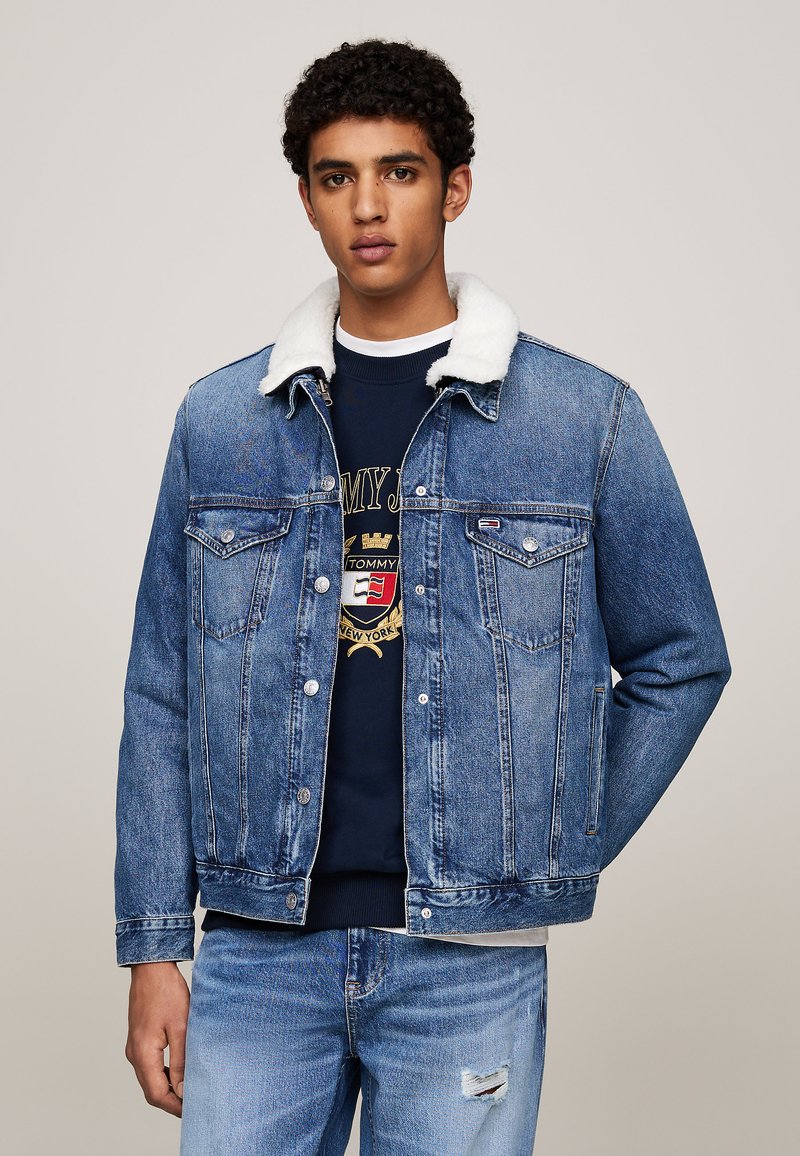 Giacca di jeans con colletto in shearling bianco, dotata di chiusure a bottoni, tasche sul petto e una lavatura azzurro chiaro; indossata sopra un maglione blu scuro.