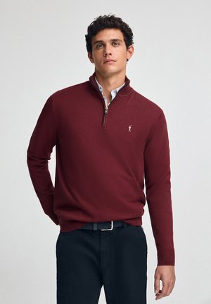 Bordeaux ritsweater met een textuurgebreide stof, voorzien van een kwart rits en een klein geborduurd logo op de linkermouw. Gecombineerd met donkere broek.