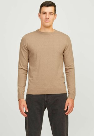 Hombre vistiendo un suéter beige de manga larga y jeans negros de pie contra un fondo liso y claro.