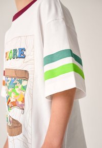 Fehér póló bordó nyakkivágással, színes "EXPLORE" grafikával, valamint zöld és kék csíkos díszítéssel az ujjain. Pamut anyagból.