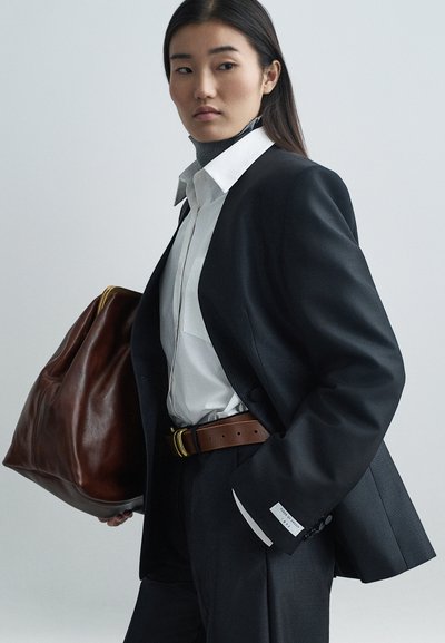 Zwarte op maat gemaakte blazer over een witte blouse, bruine leren riem met gouden gesp, en grote bruine leren tas. Strak, modern ontwerp.