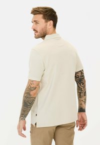 Lyse beige polo shirt, korte ærmer, ribbet krave, sideslids ved sømmen, parret med khaki bukser. Viser synlige tatoveringer på armene.