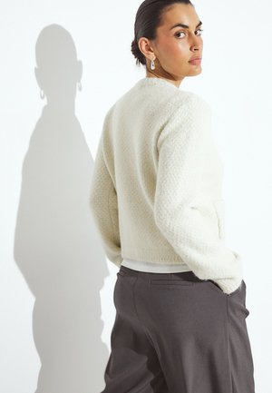 Baltas tekstūruotas megztinis su trumpa forma ir šoninėmis kišenėmis, derinamas su tamsiomis kelnėmis. Šešėlis matomas ant sienos.