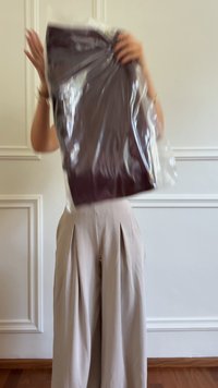 Persona che tiene un indumento scuro piegato all'interno di un'imballaggio di plastica trasparente, indossando pantaloni plissettati beige, in piedi contro una parete bianca a pannelli.