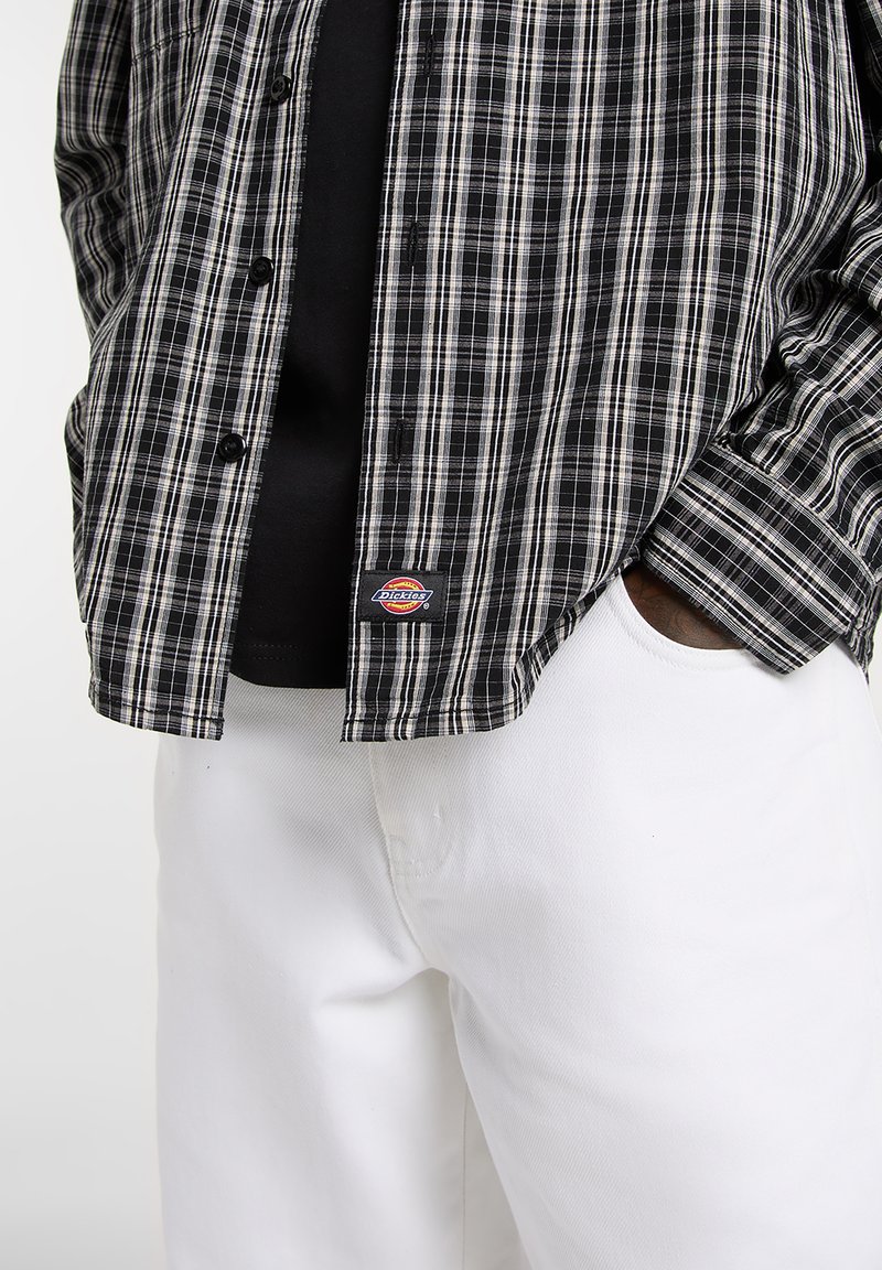 Camicia a quadri bianca e nera con design a bottoni, indossata sopra una t-shirt nera, abbinata a pantaloni bianchi. Presenta un'etichetta con il marchio sulla camicia.