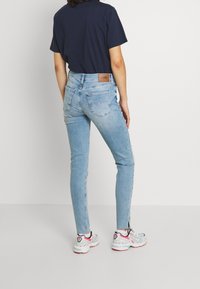 Jeans ajustados de color azul claro con una cintura alta, que cuentan con un parche de logo de cuero marrón y bolsillos traseros. Llevados con una camiseta azul marina y zapatillas deportivas.