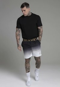 Crna majica kratkih rukava u kombinaciji s kratkim hlačama s gradijentnim prijelazom od tamne do bijele boje, s pojasom s zlatnim logotipom i bijelim tenisicama.