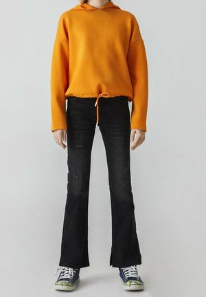 Oranssi huppari, jossa on kiristysnyöri helmoissa, yhdistettynä mustiin leveisiin farkkuihin ja sinisiin lenkkareihin, joissa on värikkäitä yksityiskohtia ja valkoiset nauhat.