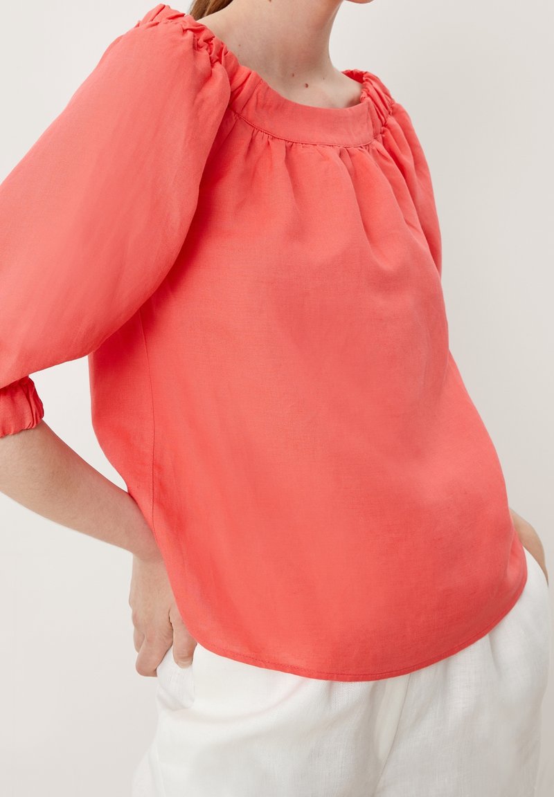 Blusa di lino corallo con maniche a sbuffo e scollatura arricciata, caratterizzata da una vestibilità comoda e una texture liscia, indossata con pantaloni bianchi.