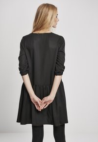 Svart kjole med tettsittende byste, full skjørt og samlet midje. Lange ermer med rullet manchetter. Glatt tekstur og enkelt design.
