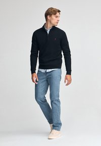 Czarny sweter na zamek błyskawiczny, lekki materiał, prążkowane mankiety i dół. Jasnoniebieska koszula pod spodem, połączona z jasnoniebieskimi dżinsami i beżowymi sneakersami.