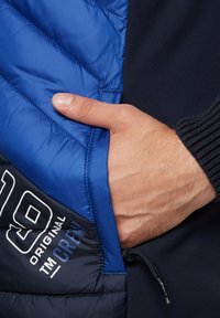 Giacca imbottita con una parte superiore blu caratterizzata da un design a chevron, corpo in un contrastante blu scuro e una mano visibile in una tasca laterale con coulisse.