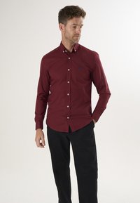 Langärmeliges, bordeauxrotes Button-Up-Hemd aus Baumwolle. Hat einen spitzen Kragen, weiße Knöpfe und ein kleines blaues Logo auf der Brust.