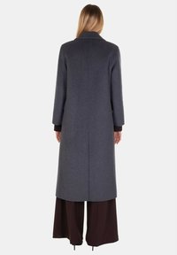 Lungo cappotto grigio in lana con colletto a revers, taglio dritto e maniche lunghe, abbinato a pantaloni svasati marrone scuro. Nessun bottone visibile.