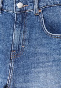 Jean en denim bleu en gros plan montrant le bouton, la braguette zippée, la boucle de ceinture et la poche avant avec un détail de couture orange visible.