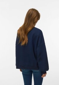 Femme aux longs cheveux roux portant un sweat-shirt ample bleu marine et un jean bleu, debout de dos sur un fond blanc uni.