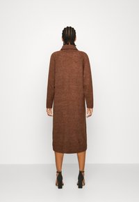 Robe longue en maille marron avec un col montant, des poignets côtelés et une coupe droite, associée à des sandales noires à talons hauts. Tissu simple et texturé.