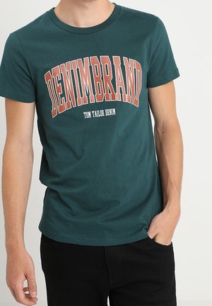 Grøn bomulds t-shirt med rund hals, prydet med fed "DENIMBRAND" tekst i orange og hvid, parret med sorte bukser.