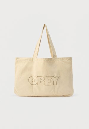 Beežist värvi kandekott kahe sangaga, mille esiküljel on suured suured tähed "OBEY" ümberjoonistatud.