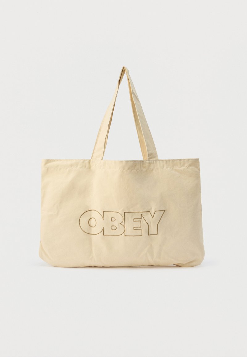 Μπεζ τσάντα ώμου με δύο λαβές και την λέξη "OBEY" με περίγραμμα σε μεγάλα κεφαλαία γράμματα στο μπροστινό μέρος.