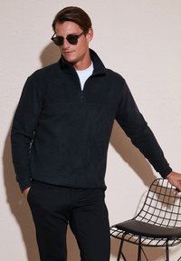 Pullover in pile nero con zip a quarto, dalla texture liscia e con un pannello a contrasto. Indossato sopra una camicia bianca e abbinato a pantaloni neri.