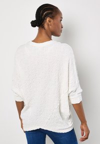 Weißer strukturierter Pullover mit Relaxed Fit, Boot-Ausschnitt und drei Viertel-Ärmeln, kombiniert mit blauen Jeans.