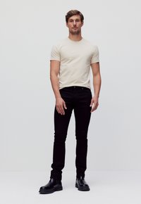 Beige katoenen t-shirt, slim fit ontwerp; zwarte slim-fit jeans; zwarte enkellaarsjes; gladde textuur, minimale details; effen achtergrond.