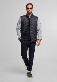 Veste hybride pour homme avec un haut matelassé sombre et des manches gris clair, dotée d'une fermeture zippée et de poches latérales. Portée avec un pantalon foncé et des chaussures.