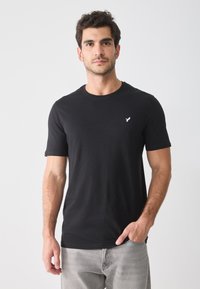 Schwarzes Baumwoll-T-Shirt mit Rundhalsausschnitt, kurzen Ärmeln und einem kleinen weißen Logo auf der linken Brust. Kombiniert mit hellgrauen Jeans.