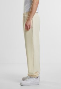 Vista lateral de una persona que lleva pantalones deportivos color crema con puños elásticos y zapatillas blancas, de pie contra un fondo blanco liso.