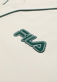 Cremefarvet stof med grøn broderet FILA-logo og matchende grøn kantdetalje nær en knap.