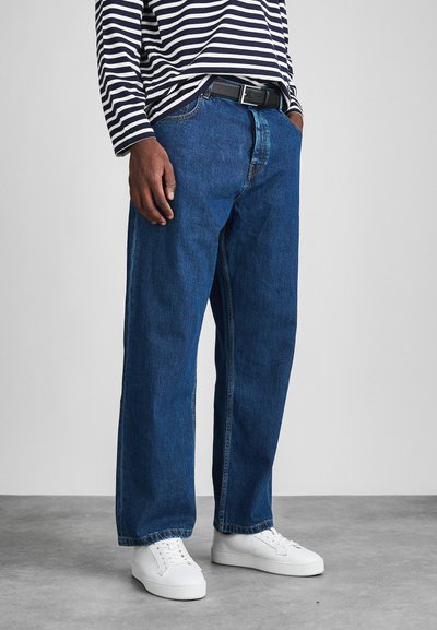 Blue denim jeans with a straight cut, five-pocket design, and visible stitching. Paired with a black belt and white sneakers.