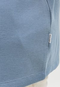 Υφασμα πλεκτό βαφτισμένο μελανζέ σε ανοιχτό γαλάζιο χρώμα με μια μικρή λευκή ετικέτα που γράφει «JACK & JONES» κοντά στη ραφή στο πλαϊνό μέρος ενός ενδύματος.