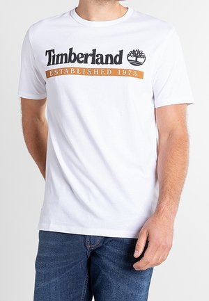 Bílé tričko Timberland s černým logem a nápisem "Established 1973" v oranžové barvě, nošené s modrými džínami na stojící osobě.