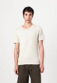 Light beige short-sleeve t-shirt made of textured fabric with a round neckline. Worn with dark, loose-fitting pants.