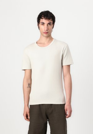 Light beige short-sleeve t-shirt made of textured fabric with a round neckline. Worn with dark, loose-fitting pants.