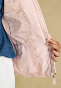 Mano che tiene aperta una giacca rosa chiaro mostrando la tasca interna e la fodera con un motivo sottile su uno sfondo beige.