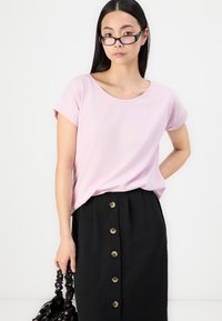 T-shirt rosa chiaro a maniche corte con collo rotondo abbinato a una gonna nera con bottoni dorati, tessuto lavorato e una borsa nera.