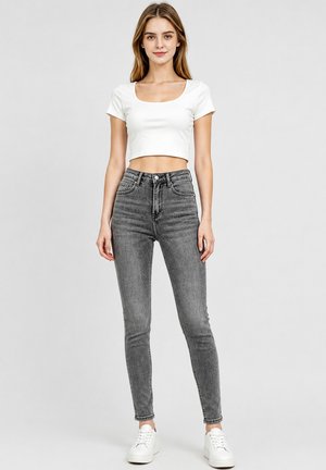 Ung kvinde står iført hvid cropped top, høj taljede grå skinny jeans og hvide sneakers mod en ensartet lys baggrund.